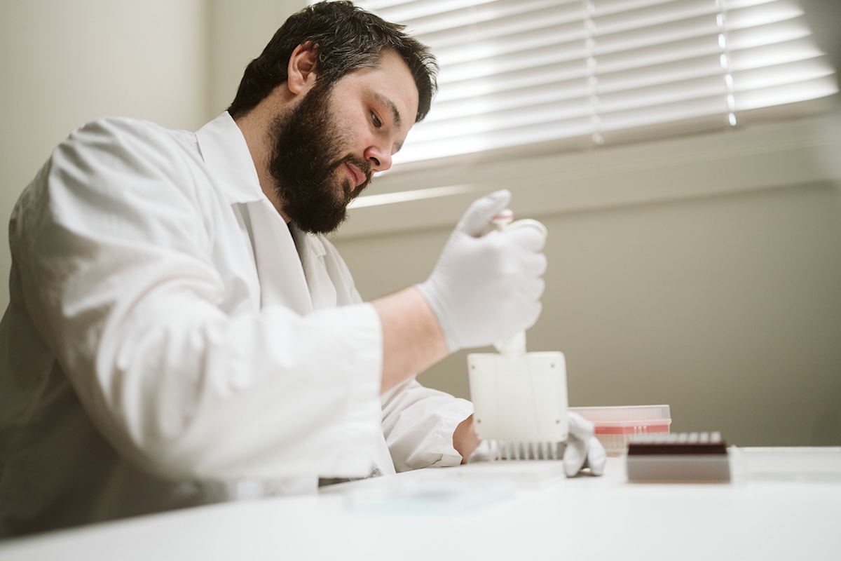 A man with light skin, dark short hair, a dark beard and a white coat and gloves prepares a sample in a white lab.