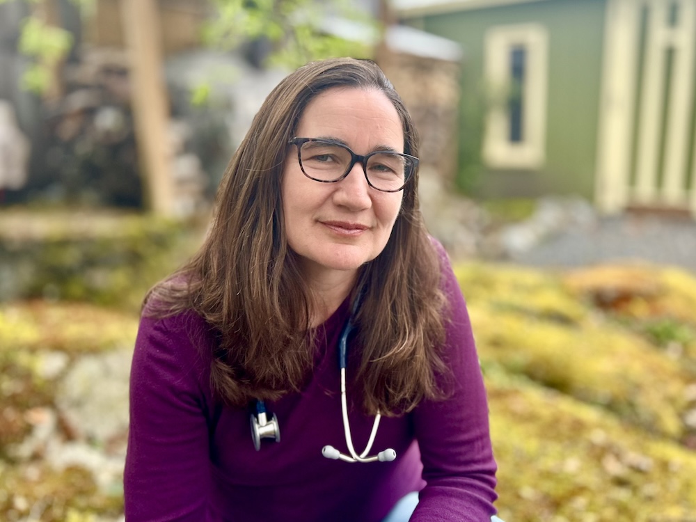 A portrait of Dr. Jill, a middle-aged woman with light skin tone and brown hair. She is wearing a purple sweater, glasses and a stethoscope.