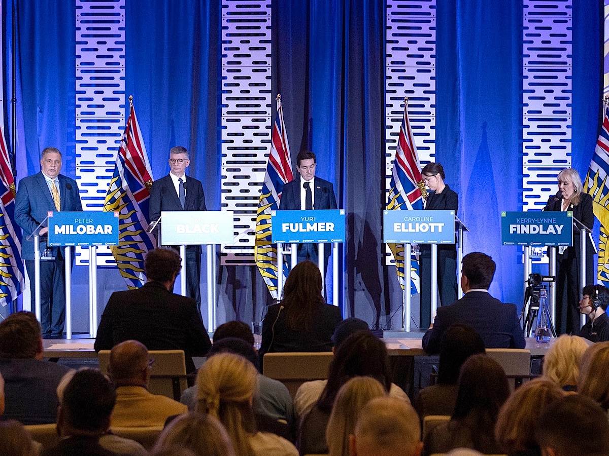 Five people, three men and two women, are at podiums on a debate set with a blue backdrop.