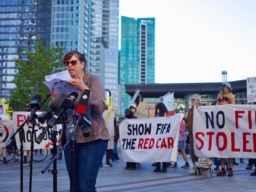 A woman wearing sunglasses speaks at microphones. Behind her people carry banners that read, &lsquo;Show FIFA the red card.&rsquo;