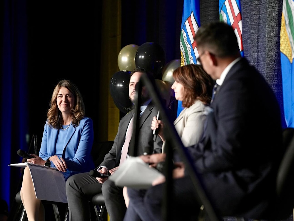 A woman with long brown hair wearing a blue skirt and blazer smiles onstage at a conference. Three other people are sitting to her right.