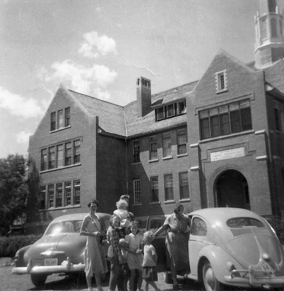 A black and white photo shows a family with parents and young children in front of a 1950s car and a brick building.