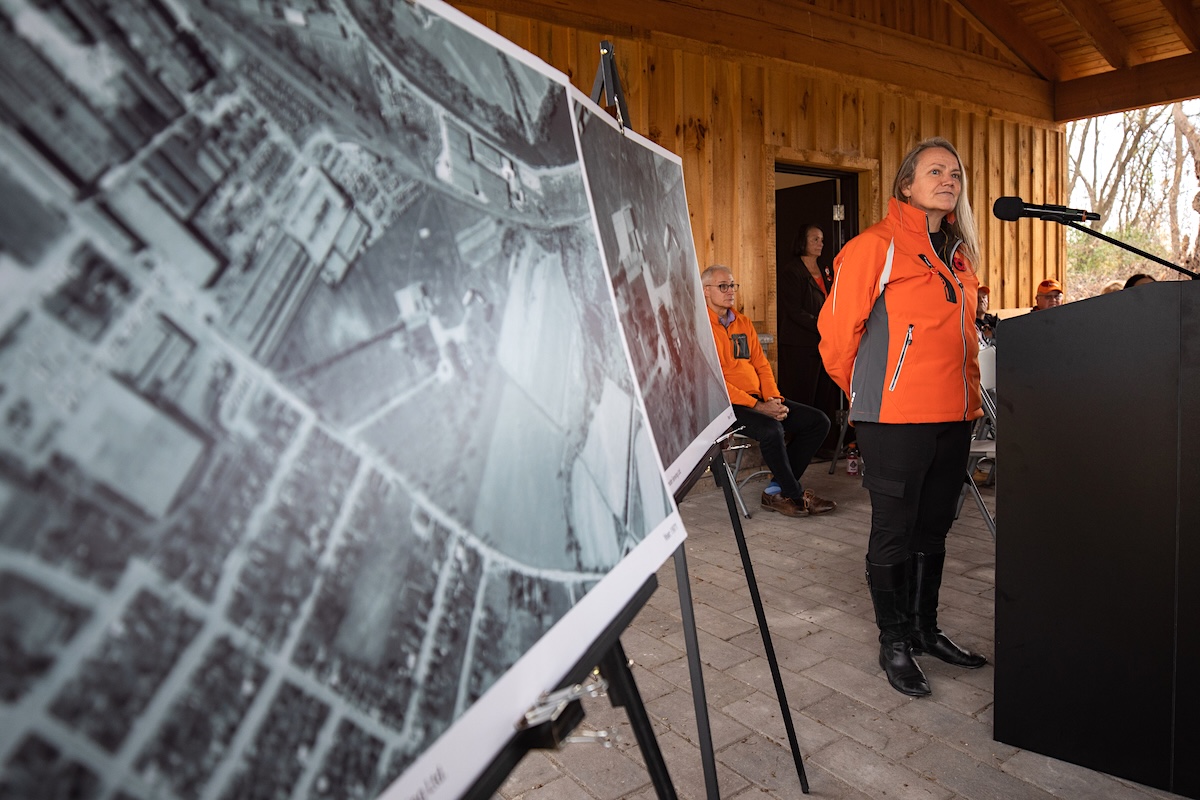 A woman wearing an orange jacket stands at a podium. To the left of the frame, an aerial photo showing some plots of land can be seen.