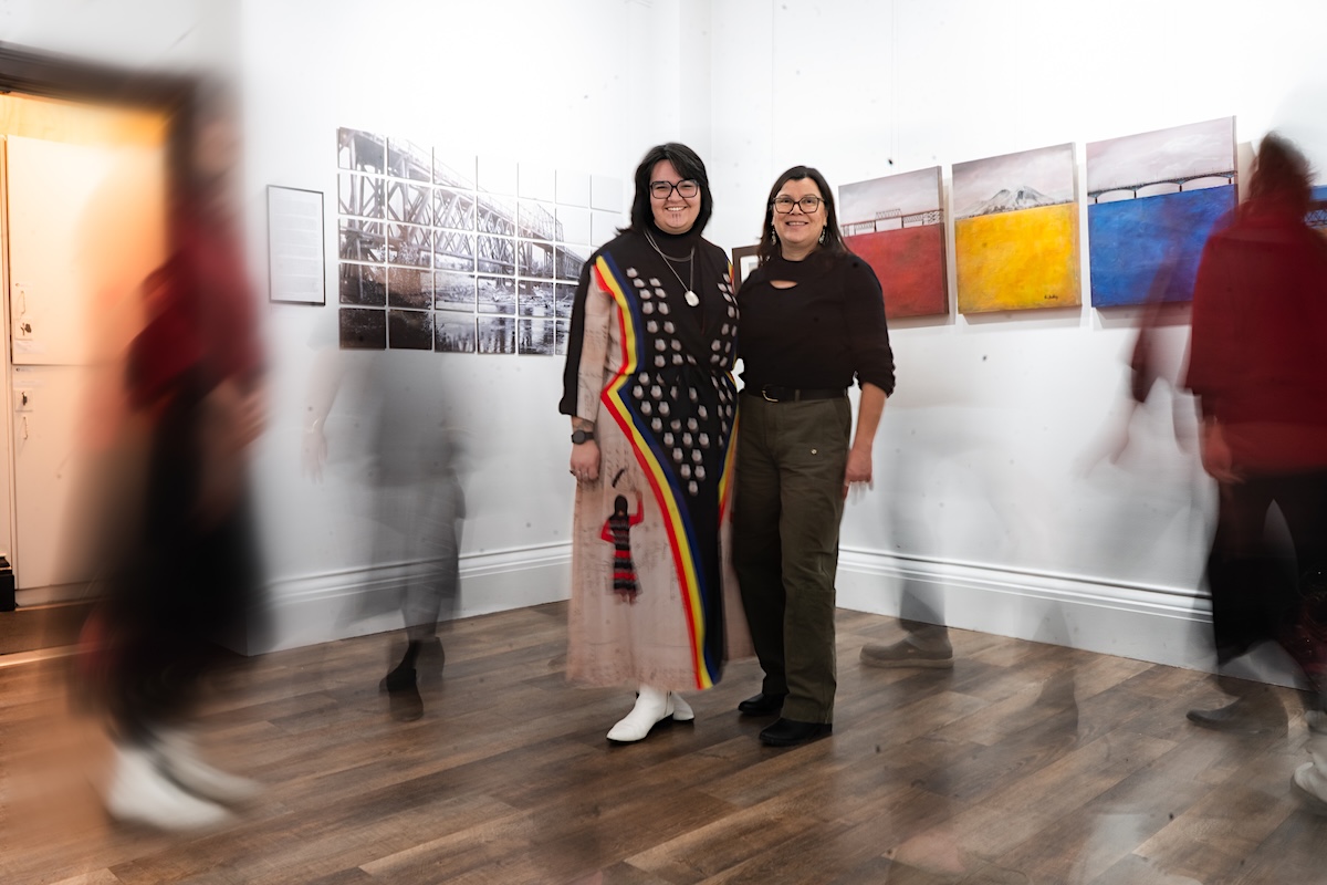 Two women with medium-light skin and dark hair stand among artwork in a gallery, surrounded by blurred figures in motion.