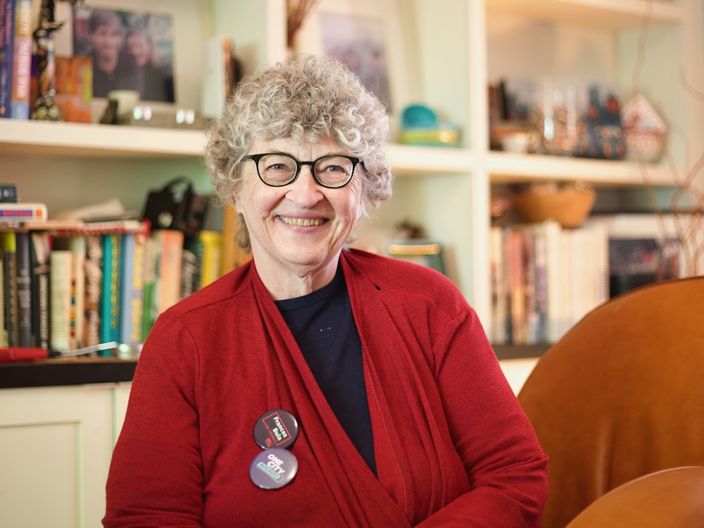 A woman in her 70s with curly grey hair smiles at the camera. She sits in front of a bookshelf.