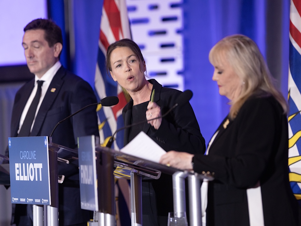 Three people stand at podiums in front of a blue backdrop and flags as they participate in a leadership debate.