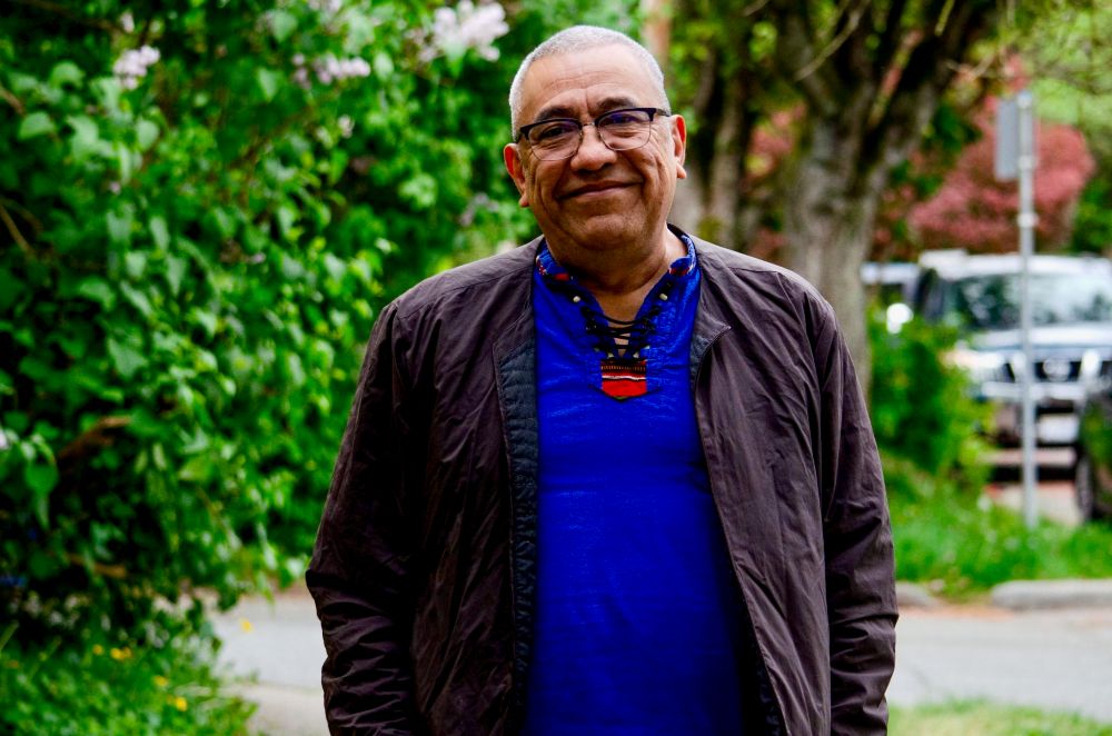 A man in his 60s stands on a tree-lined street. He has short salt-and-pepper hair and wears glasses.