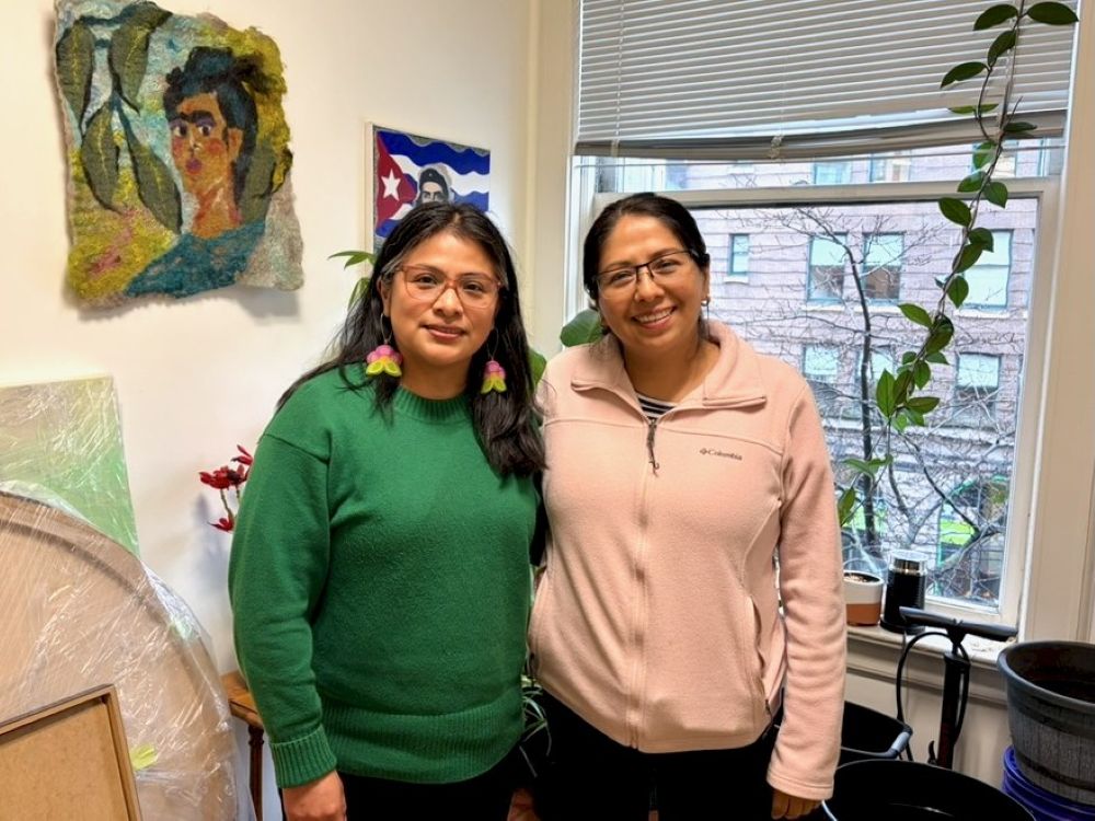 Two women stand in a room with a window behind them. The woman on the left has long black hair and wears glasses and a green sweater. The woman on the right has her hair pulled back and wears a light pink fleece jacket.