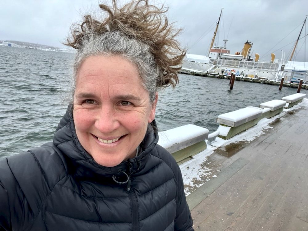 A woman stands on a dock with boats in the background. She has long gray and brown hair pulled into a ponytail.