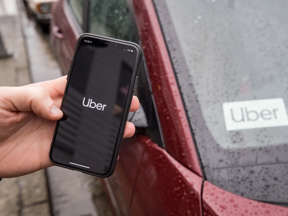 A person&rsquo;s hand holds a phone with the Uber logo on the screen, in front of a red car with an Uber logo displayed on the dashboard.