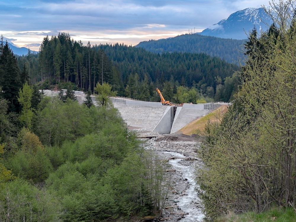 A large, dam-like barrier sits in a mountain valley. There is a gap in the middle that allows a rushing mountain river to flow through it. 