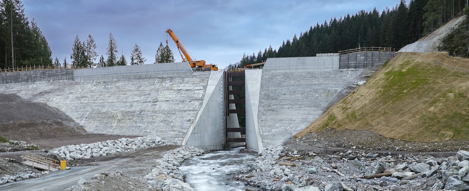 A piece of heavy equipment can be seen atop a dam-like structure with a narrow gap that allows river water to pass through.