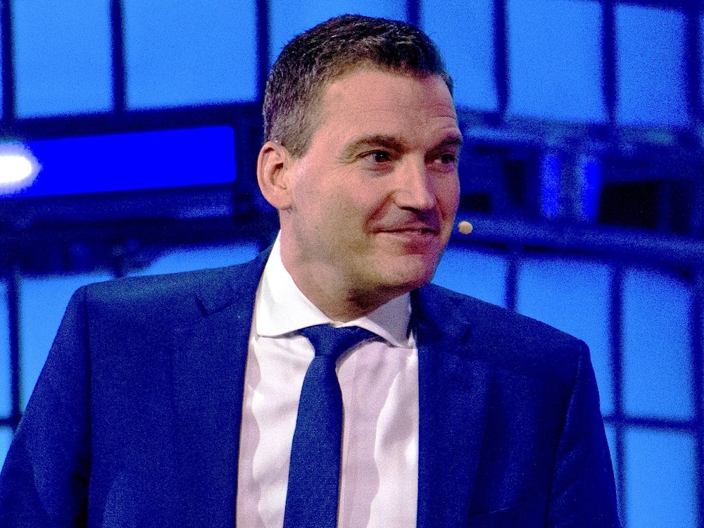 A man with short dark hair wearing a dark blue suit sits onstage in front of a glowing blue background.