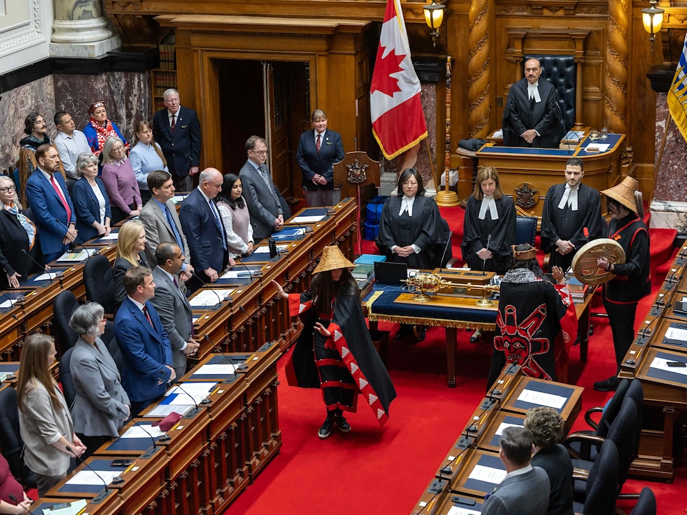 The photo looks down in the B.C. legislature where three Indigenous people in regalia stand between rows of MLAs.