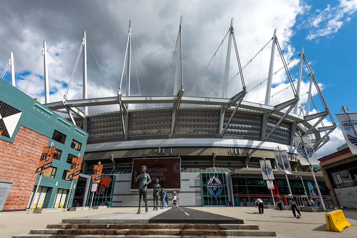 A few people walk around BC Place in the daytime. Whitecaps flags are up.