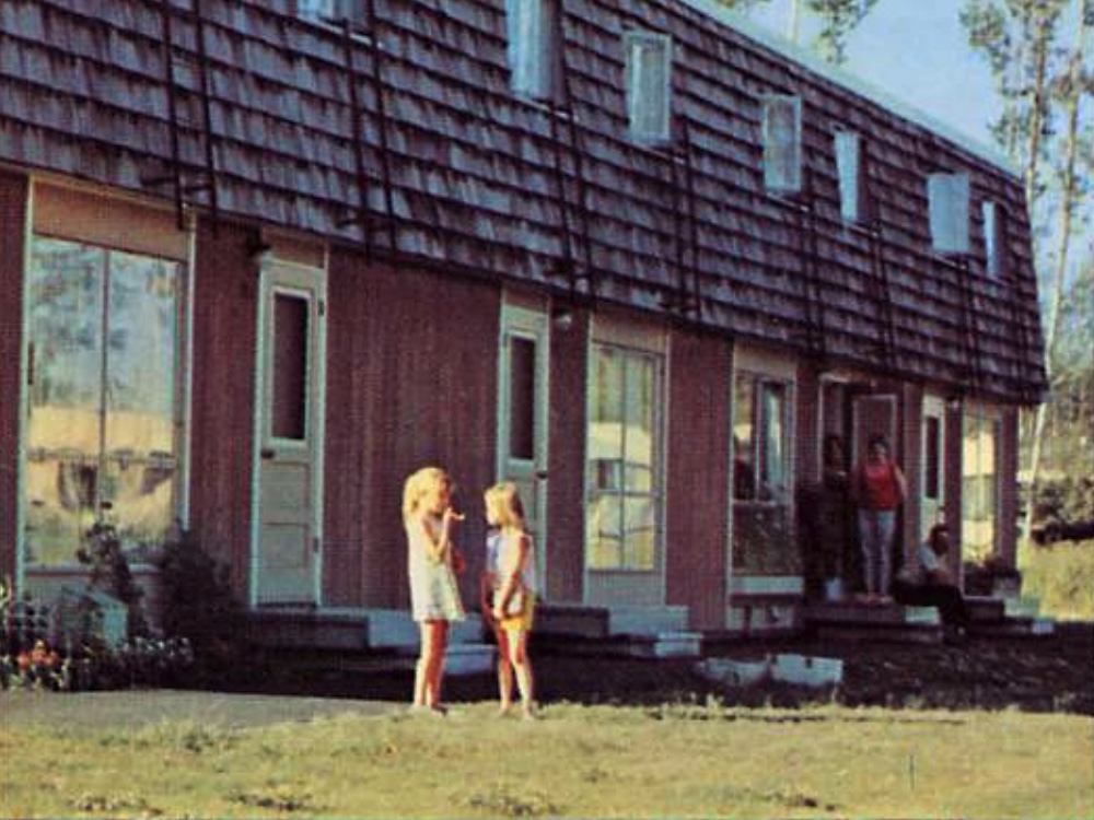 Two young children stand on a lawn outside a group of row houses.