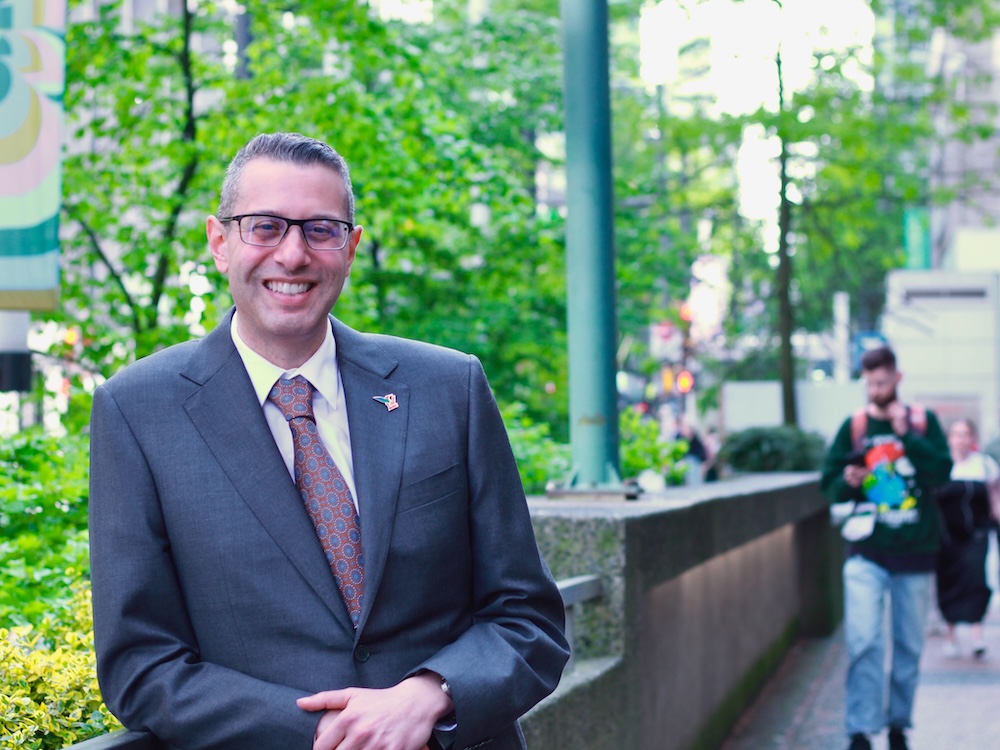 A middle-aged man with glasses and curly salt-and-pepper hair stands on a downtown street. He leans on a railing and wears a blue suit.