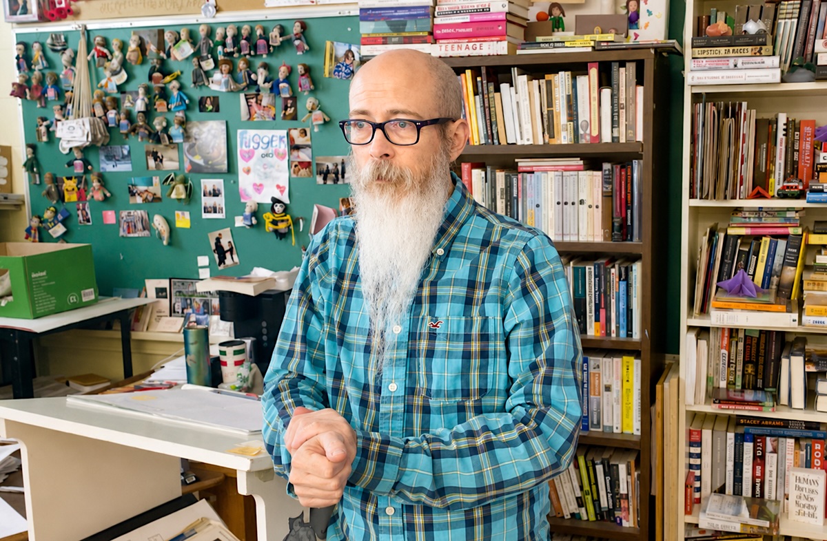 A man with light skin tone and a long grey beard wears glasses and a blue plaid shirt. He is standing in a decorated classroom with books and literary figurine magnets.