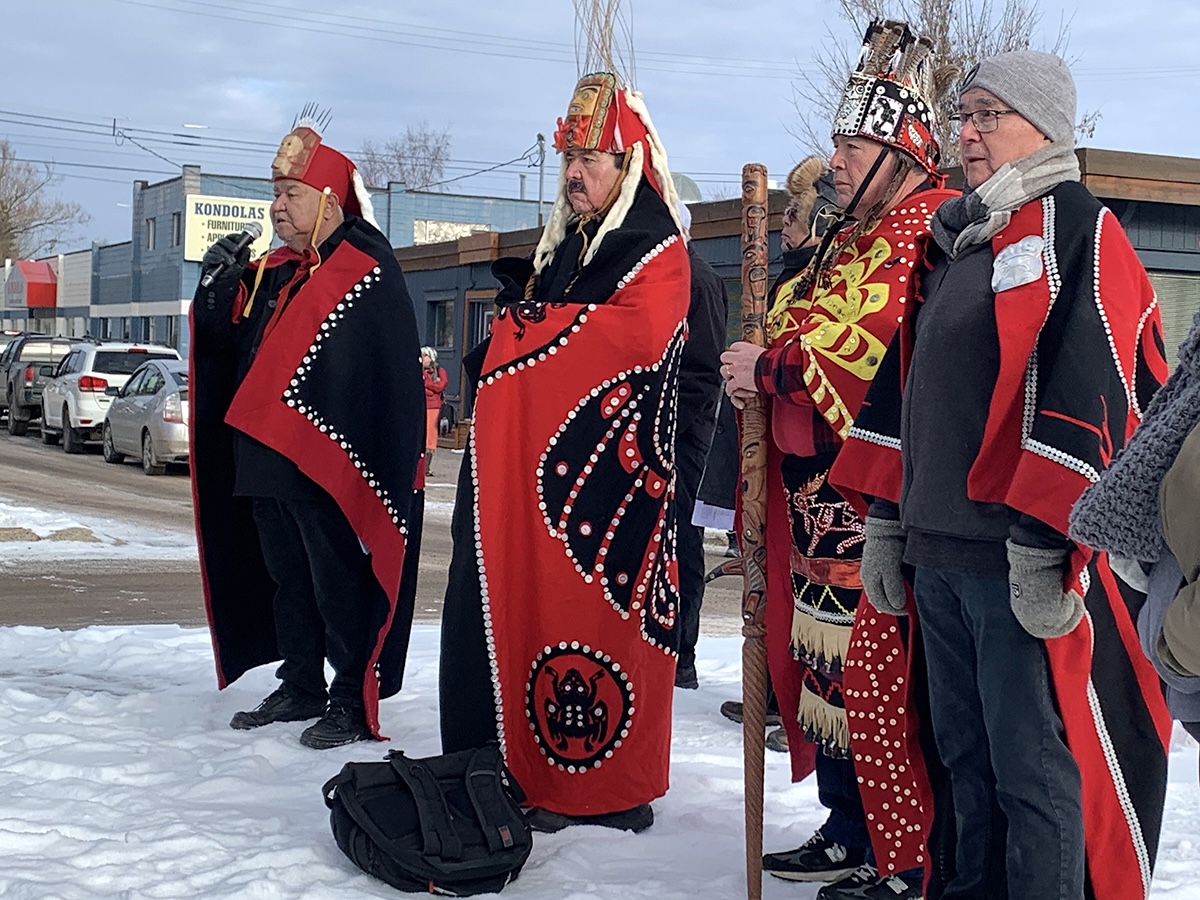 Four men in traditional First Nations regalia stand in a snowy setting. One speaks into a microphone.