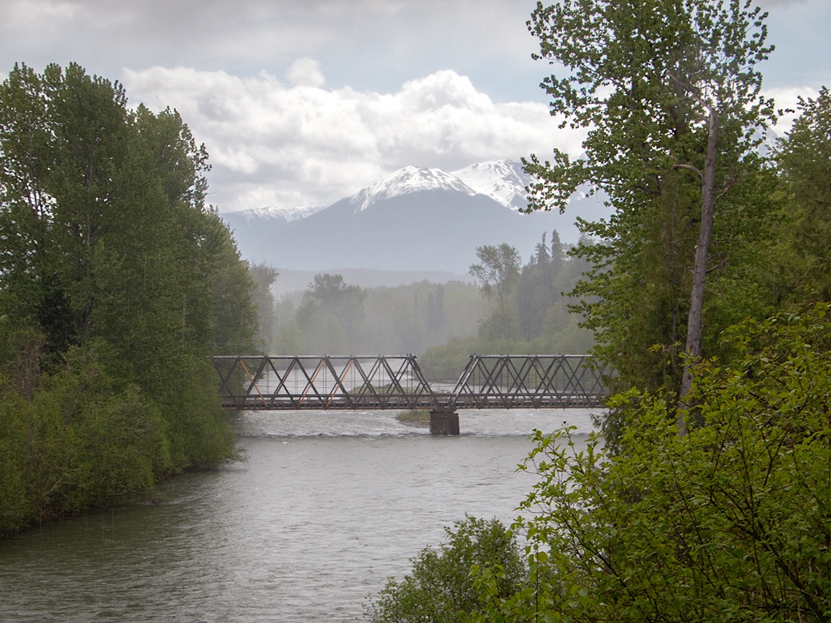 A scenic photo of a bridge over a river in a forested setting with mountains beyond.