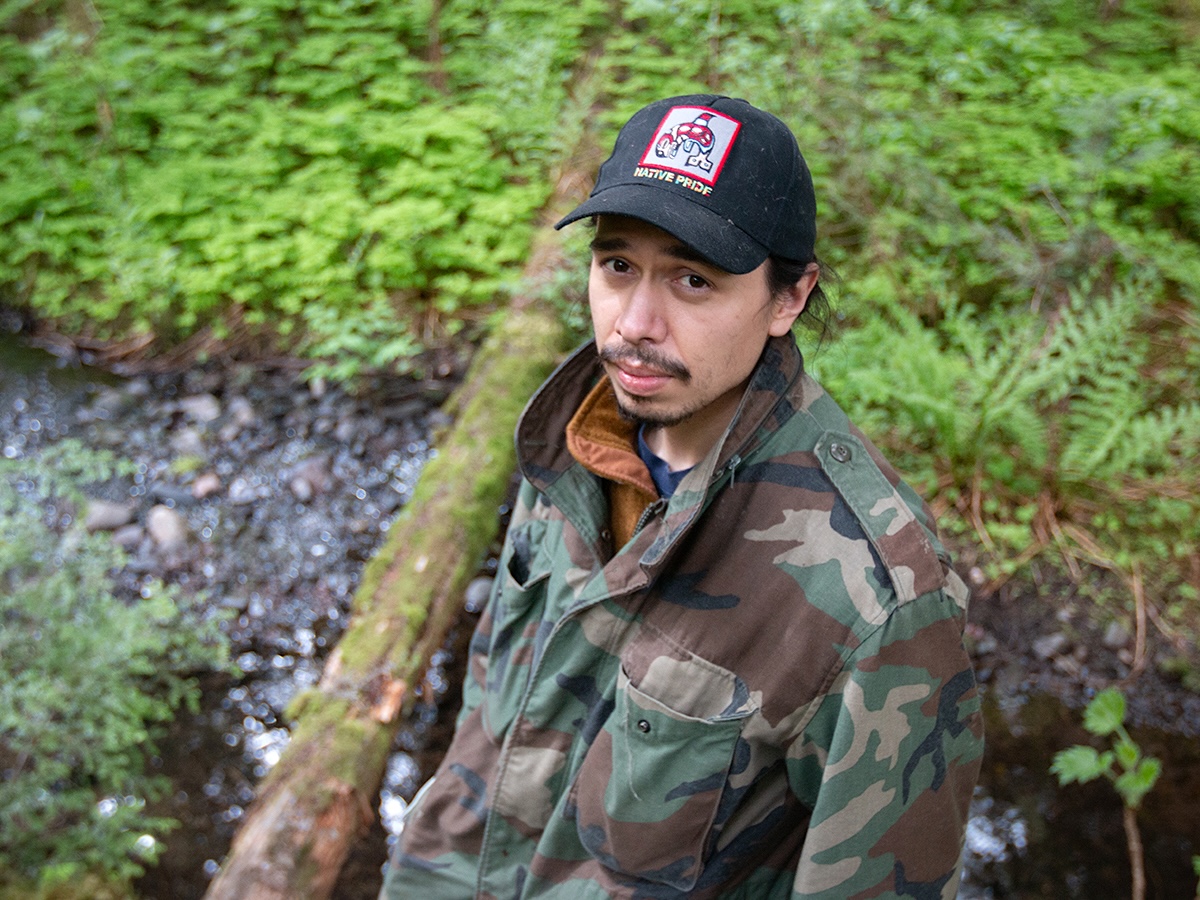 A man with dark hair and skin tone, wearing a camouflage jacket and black ball cap that says &lsquo;Native pride,&rsquo; stands in a wooded landscape.