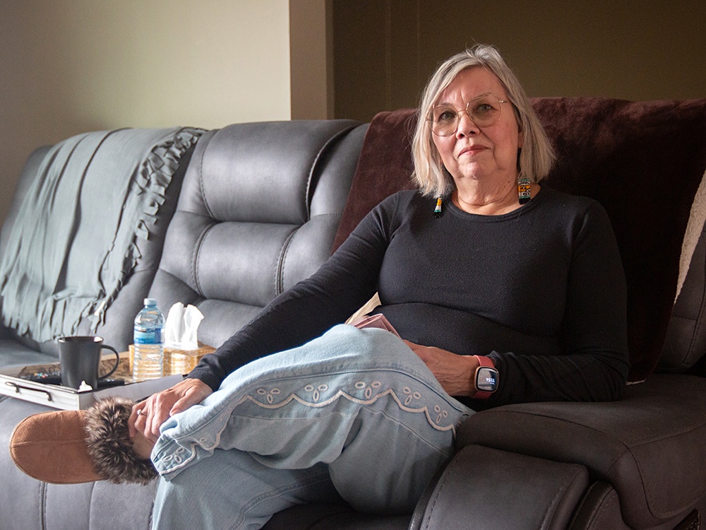 A woman with shoulder-length hair and glasses sits on a couch in her living room. 