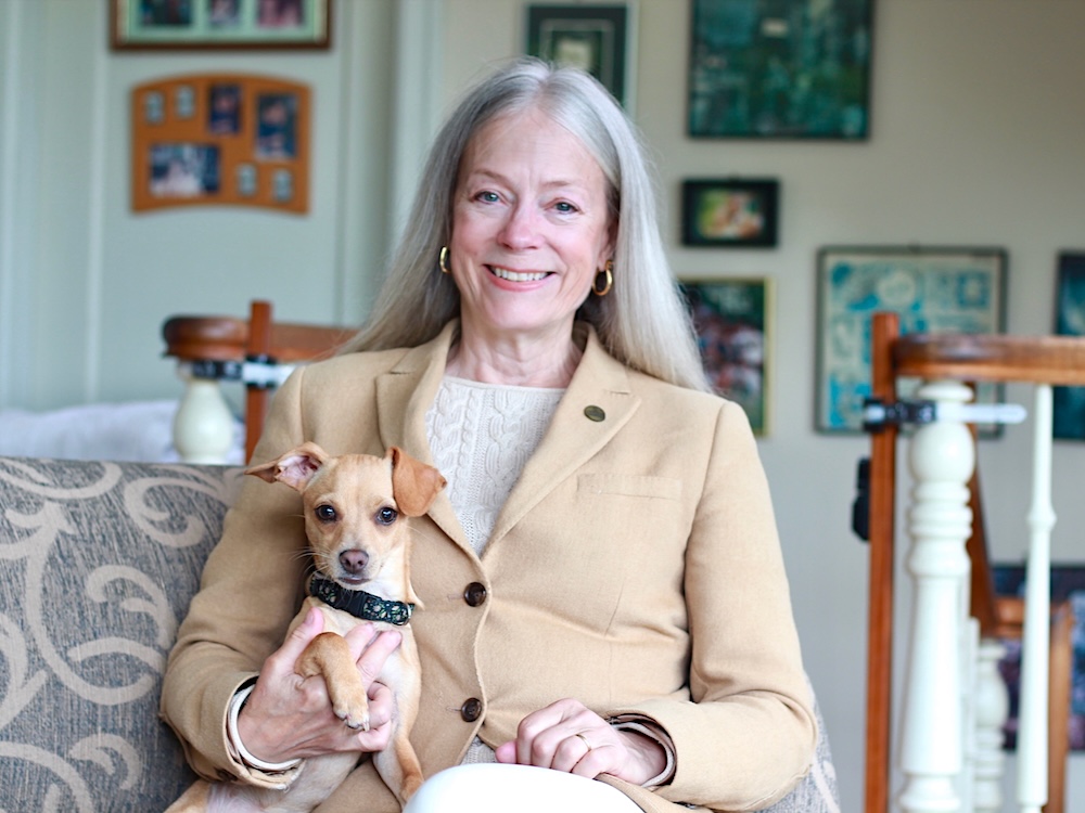 A woman with light skin and long grey hair wears a tan jacket in a cosy living room setting. She holds a small dog in her right arm.