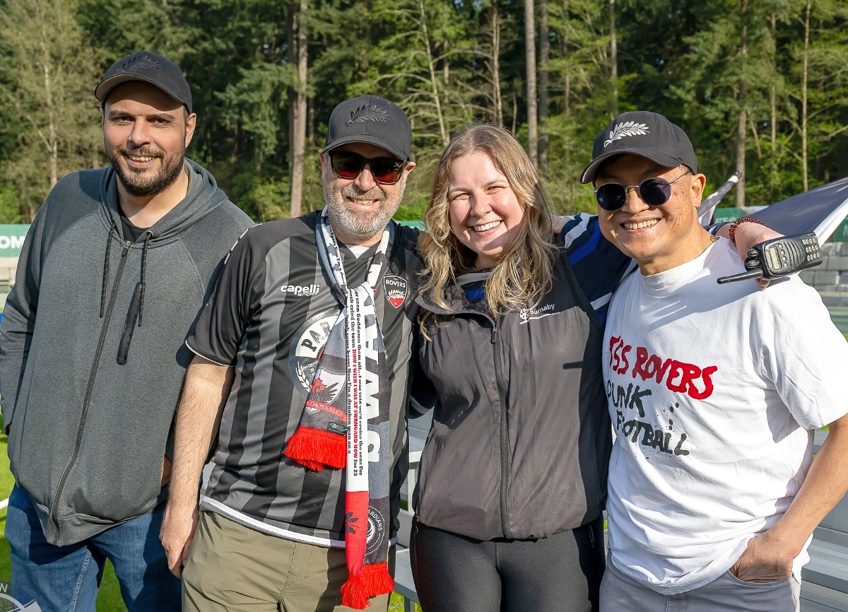 Three men and a woman in various casual attire smile and pose for a photo on a sunny day.