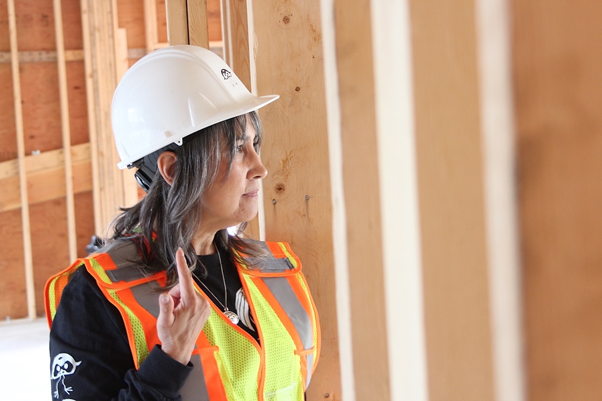 A woman with medium-light skin tone and shoulder-length grey hair wears a high-vis vest and a hard hat and stands in a building under construction.