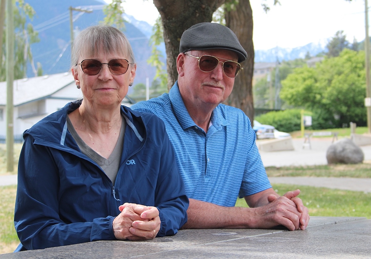 An older woman and man with light skin tone and grey hair wear sunglasses and sit at a picnic table.