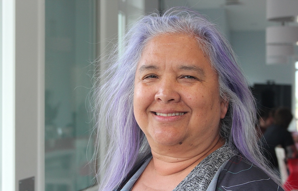 A woman with medium-light skin tone and long purple-grey hair smiles at the camera.