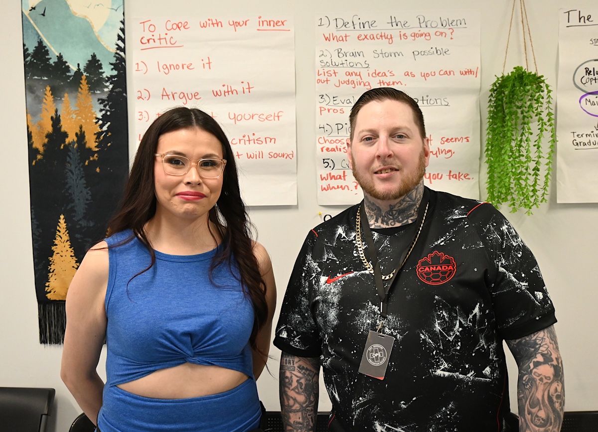 A woman with black hair, glasses and a blue dress stands next to a man with tattoos and a black and white patterned T-shirt. They smile in a friendly way. 