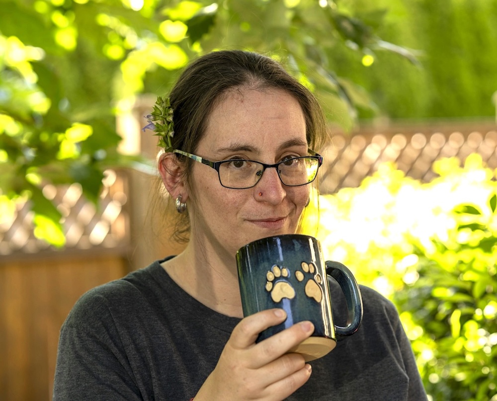 A woman with tied-back brown hair, light skin, glasses and a nose stud holds a mug with two paw prints on it. She is outdoors in a sunny fenced yard.