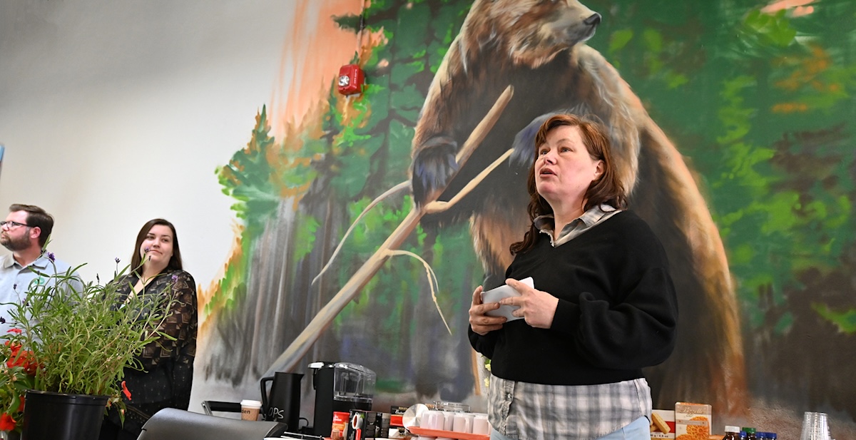 A woman with brown hair and a black sweater stands in front of a mural of a large bear.
