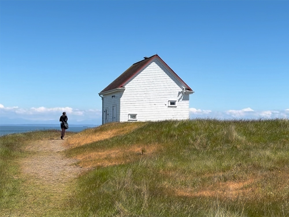 A small white building with wooden siding can be seen against a blue sky, the ocean and a windswept, grassy area. There is a dirt path and one person on a bicycle.