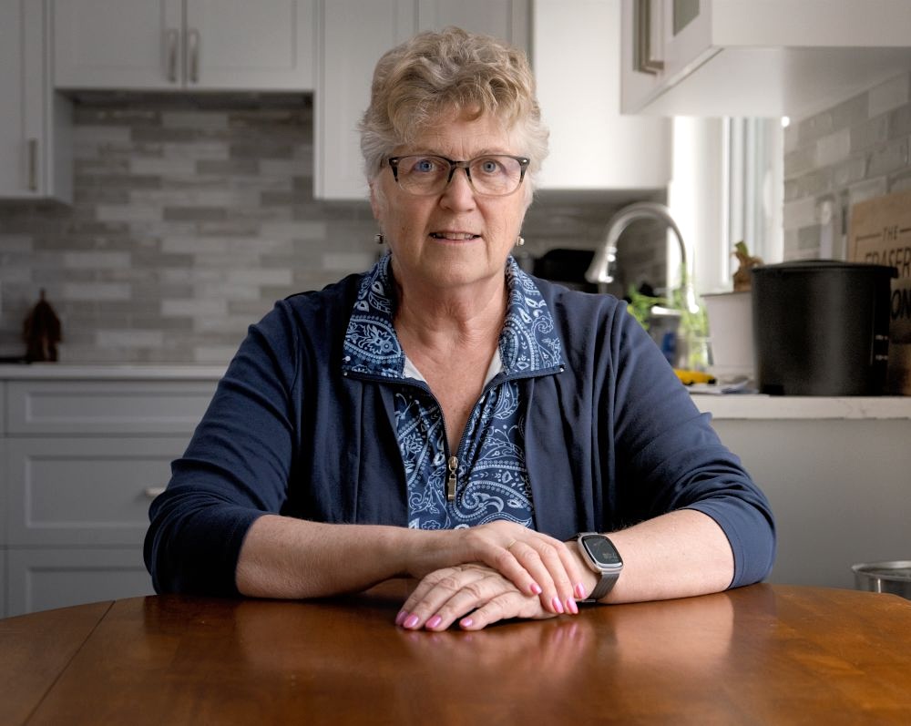 A woman with grey-blond hair and glasses sits at a table in a kitchen.