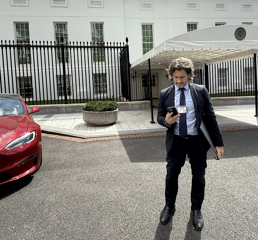Barron, on the right, looks at his phone. He&rsquo;s in front of a canopied walkway leading to the West Wing. A red car, to the left, is partially in frame.