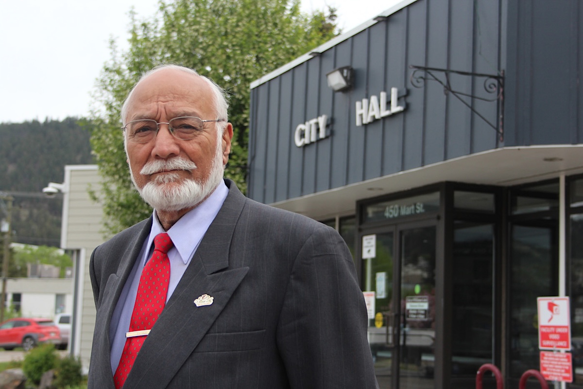 A balding man with medium-light skin tone, white hair and a beard wears a suit and stands in front of a single-storey building that says &lsquo;City Hall.&rsquo;