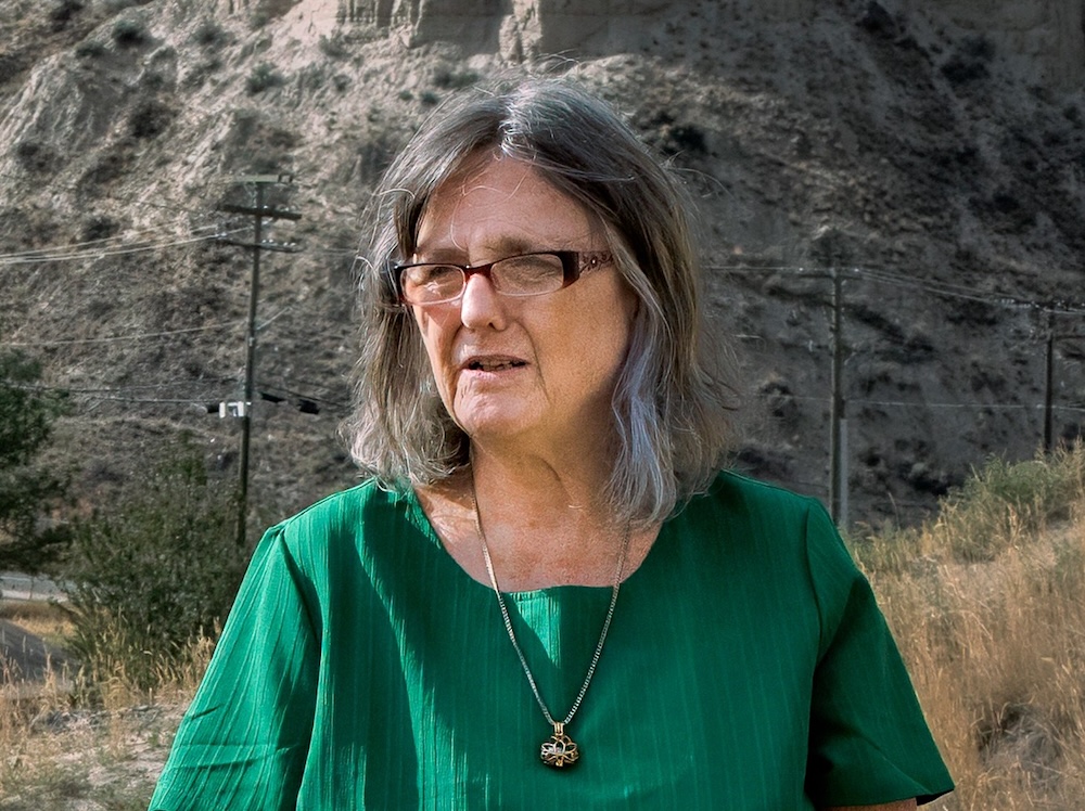 Dale Bass, a woman with shoulder-length grey hair and glasses, wearing a green top, stands in an arid landscape.
