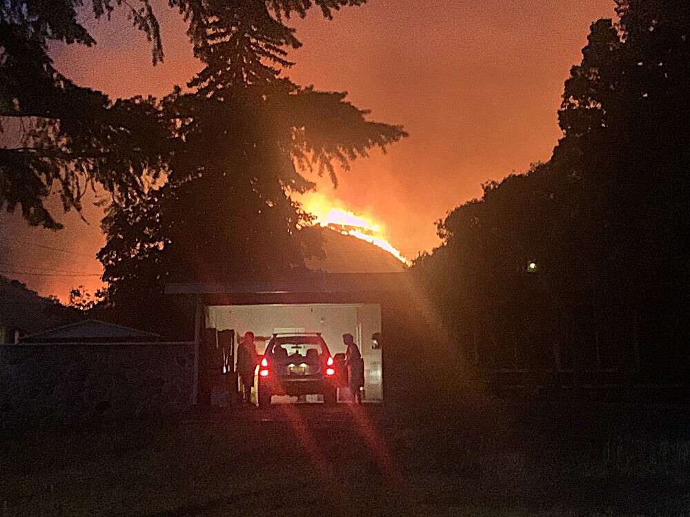 At night, with a hillside burning behind a house, an open garage door reveals the silhouettes of people getting into a car.