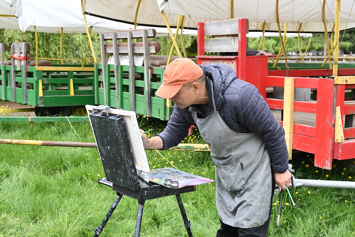 A man in an orange baseball hat and blue jacket with a grey smock bends over an easel. He is standing in front of brightly coloured wagons and is holding several paintbrushes in his hands. 