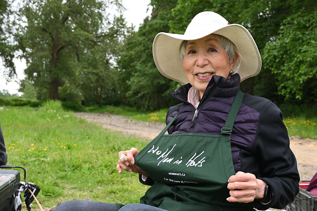 A woman with grey short hair and a white hat holds out her modified smock. It reads &lsquo;Men in Hats,&rsquo; but she&rsquo;s added to it so it reads &lsquo;Women in hats.&rsquo;