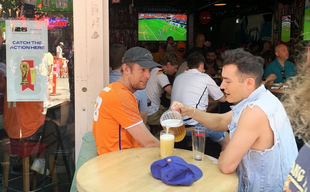A man in a sleeveless denim top pours beer into his glass from a pitcher while sitting beside a man in an orange jersey at a downtown Vancouver bar.