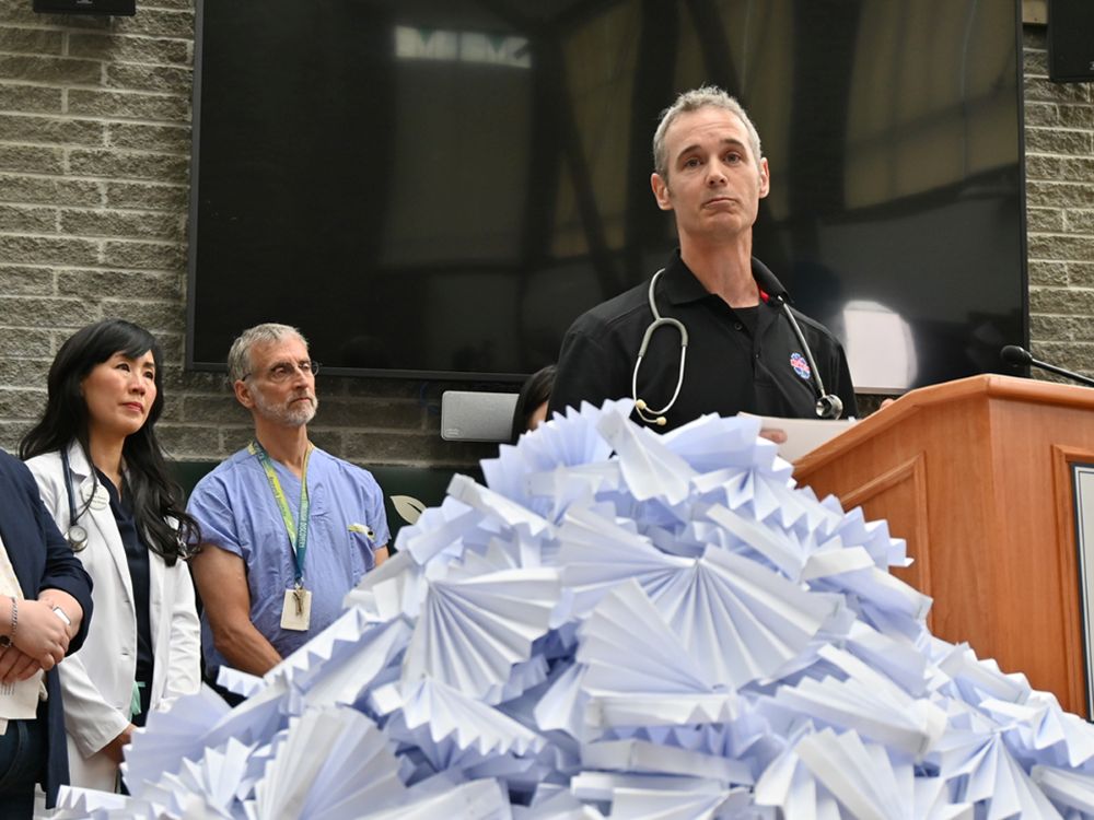 A man with short light hair stands in front of a black television screen. He is wearing a black uniform and has a stethoscope around his neck. There is a large pile of white fans in the foreground. Organizers for the event made 619 fans to represent the 619 British Columbians killed during the 2021 heat dome.