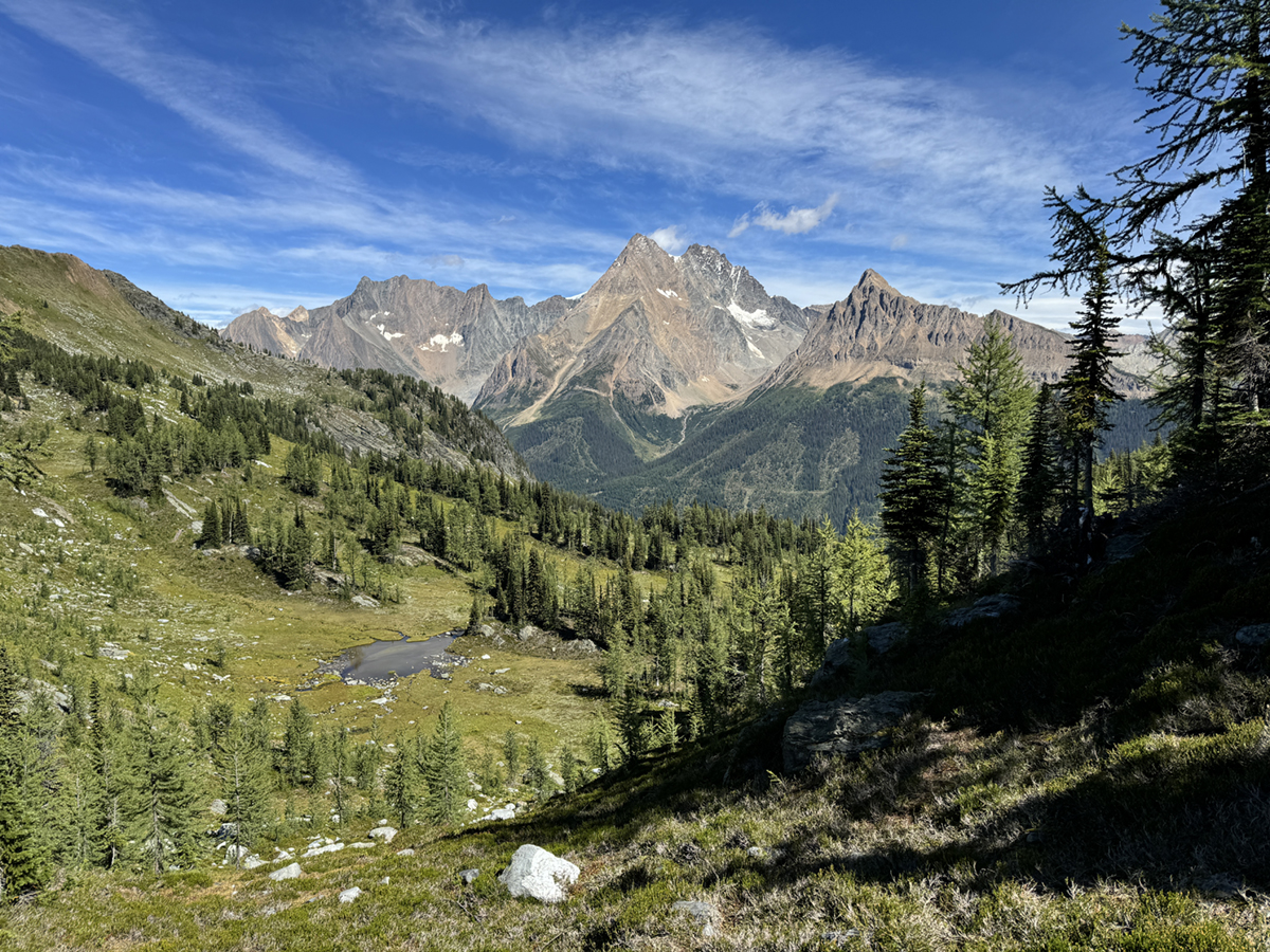 In the foreground, a small lake and a green valley. In the background, a steep, rocky mountain range. 