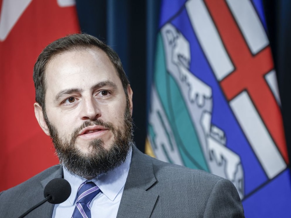 A bearded, brown haired middle-aged white man speaks with the Alberta flag out of focus behind him.