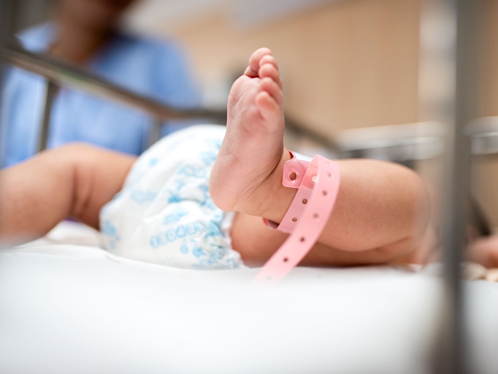 An infant lies on their back. They&rsquo;re wearing a white diaper and a pink anklet for ID. A person in scrubs stands behind them.