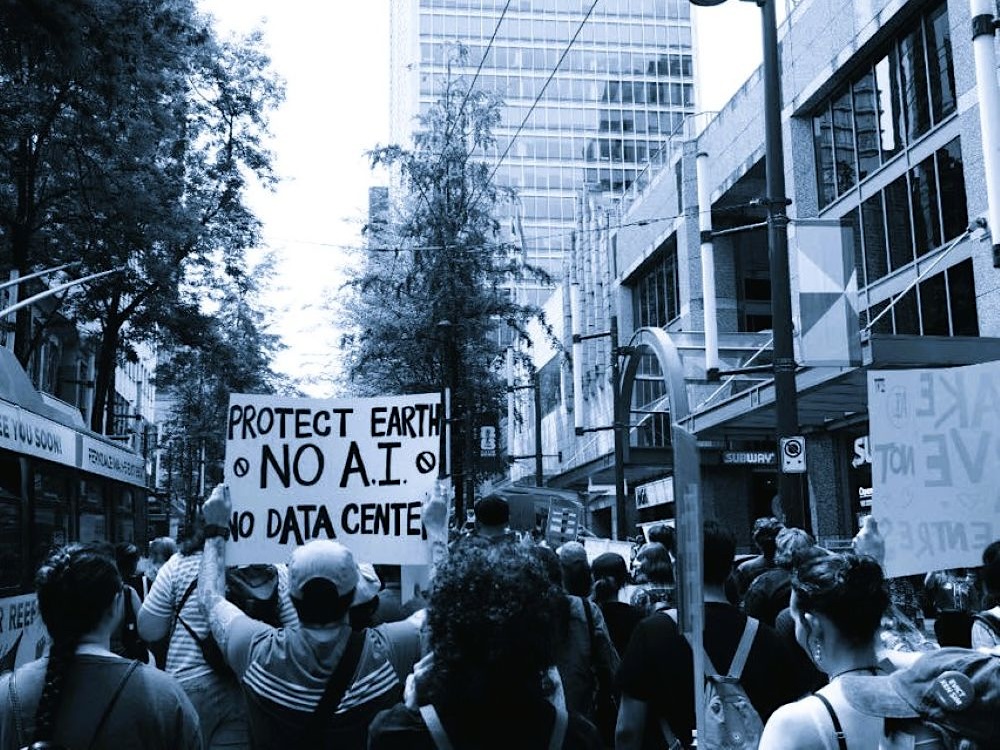 A black and white photo of a group of protesters from behind. One holds up a sign that says, &lsquo;Protect Earth. No AI. No Data Centre.&rsquo;
