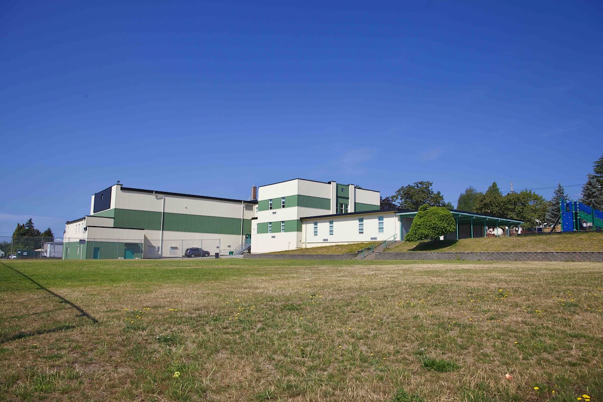 In the foreground, a grass sports field leading up to a green and white building that is an elementary school against a blue sky.