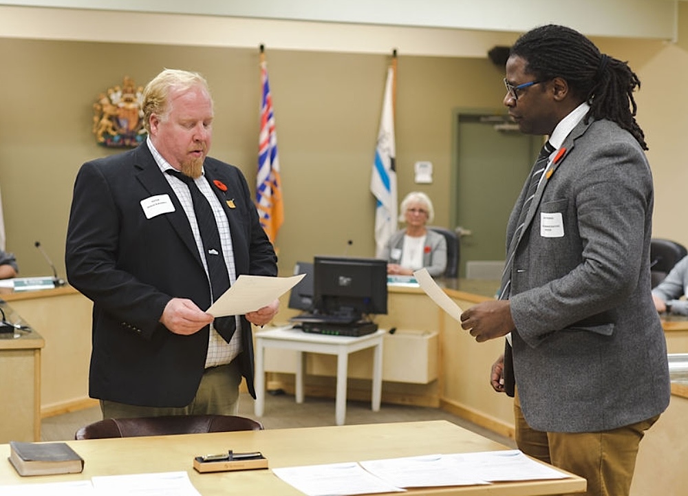 A light-skinned man with a goatee and wearing a suit reads from a piece of paper in a small community&rsquo;s council chambers.
